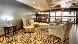 A lobby with a patterned carpet, seating area with sofas and armchairs, a side table with a lamp, and a bookshelf.