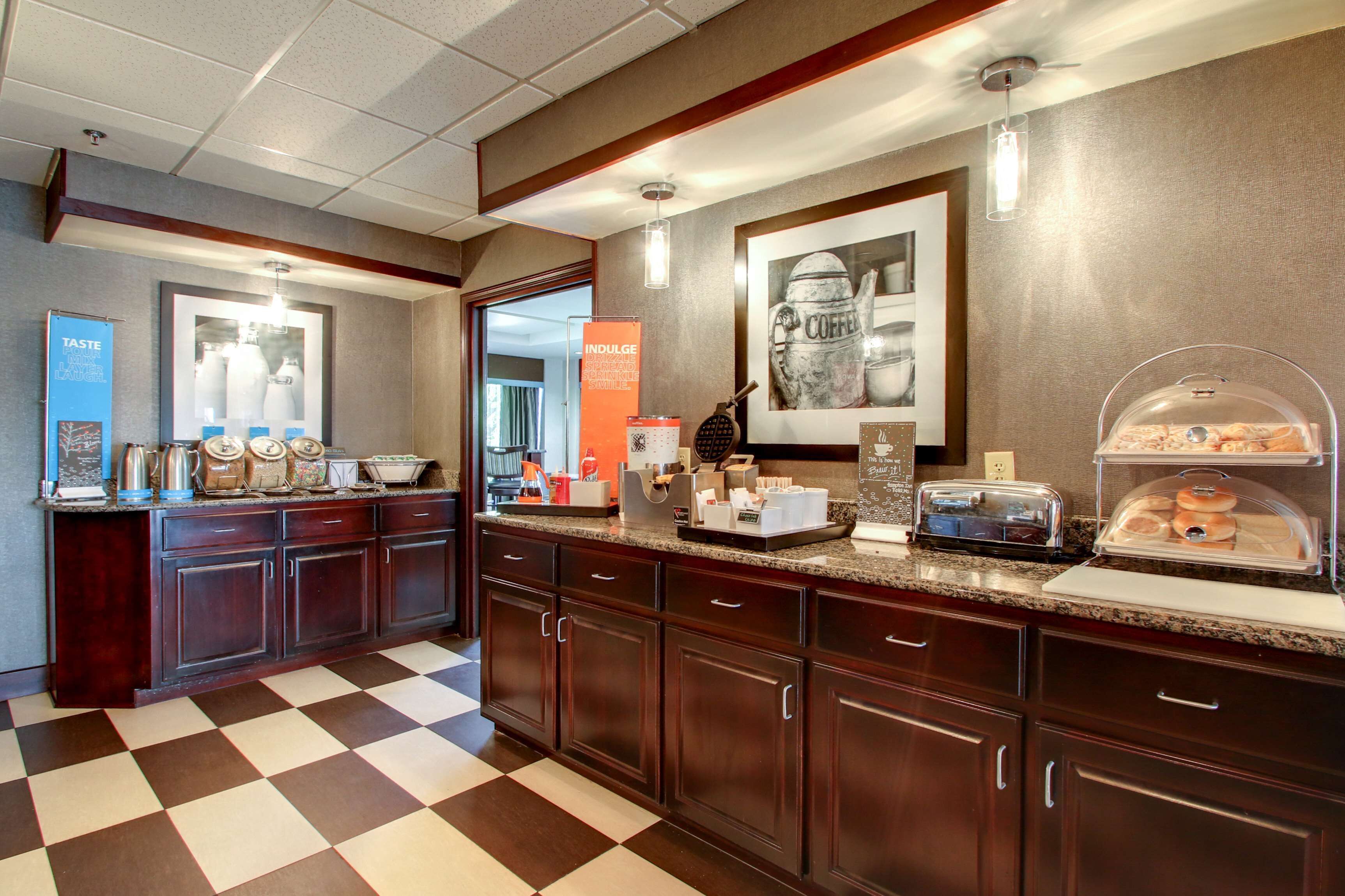 A hotel kitchen with a checkered floor, wooden cabinets, and various breakfast items on the counter.