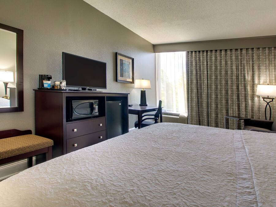 Ein Hotelzimmer mit einem großen Bett, einem Schreibtisch mit Stuhl, einem Fernseher, einer Mikrowelle und einem Fenster mit Vorhängen.