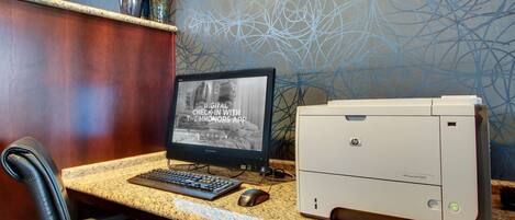 A hotel desk with a desktop computer, a printer, and a monitor displaying a digital check-in message.