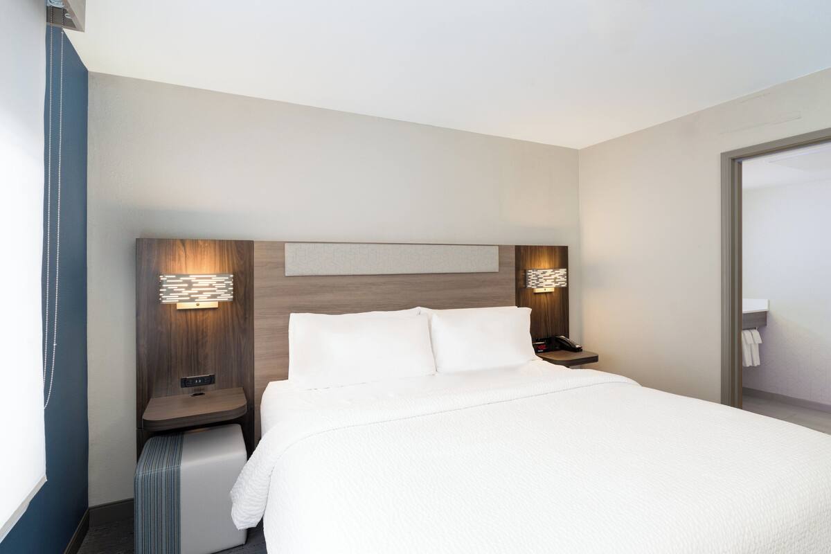 A modern hotel room with a large bed, bedside tables, a nightstand, and a wall-mounted light fixture.