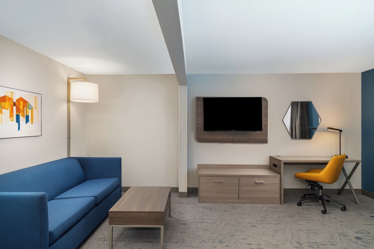 Quarto de hotel moderno com sofá azul, escrivaninha de madeira, cadeira de escritório amarela, TV de tela plana e um quadro na parede.