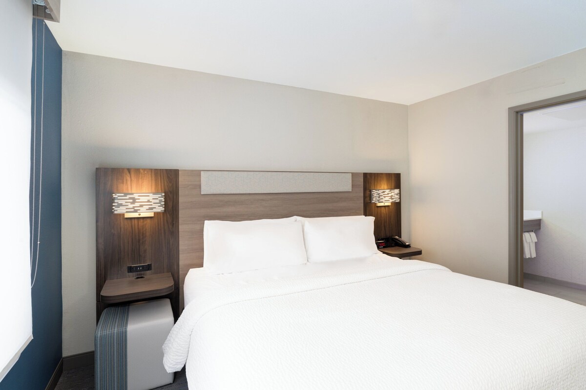 Quarto de hotel moderno com uma cama grande, mesas de cabeceira e luminária fixada na parede.