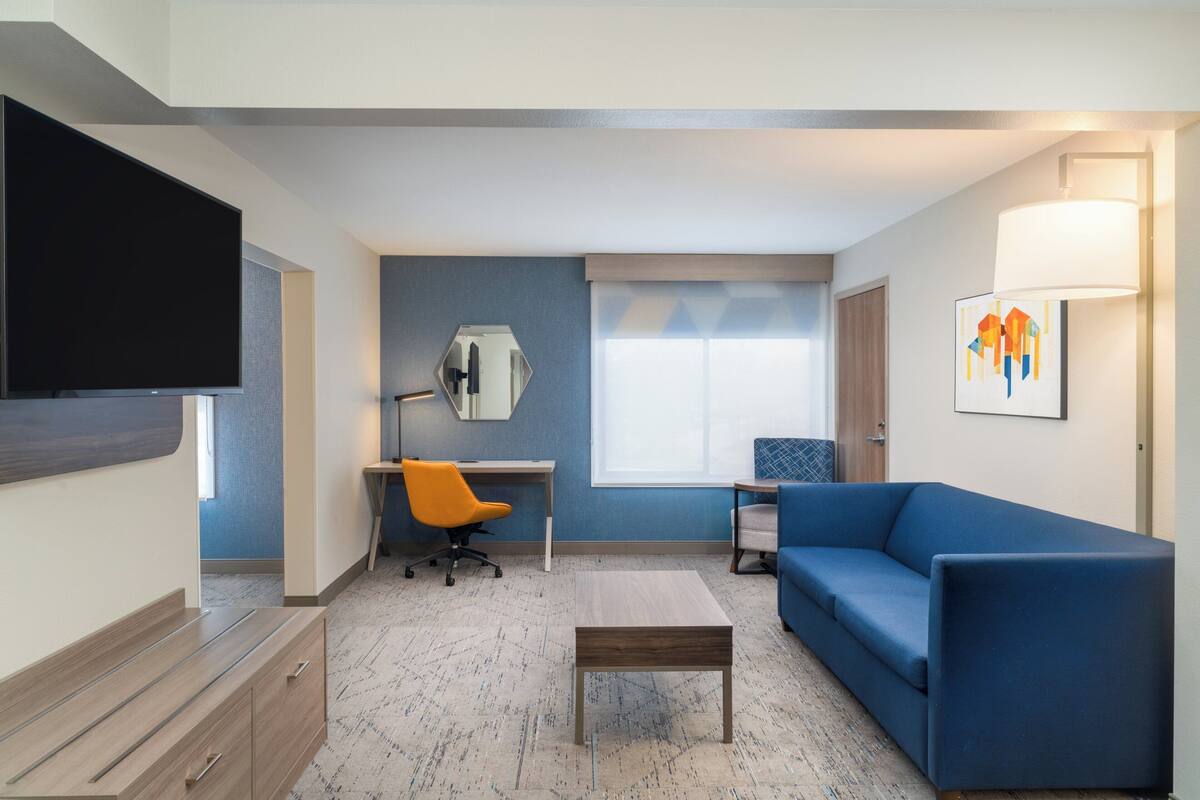 A modern living room with a flat-screen TV, a blue sofa, a wooden coffee table, an orange office chair, and a desk.