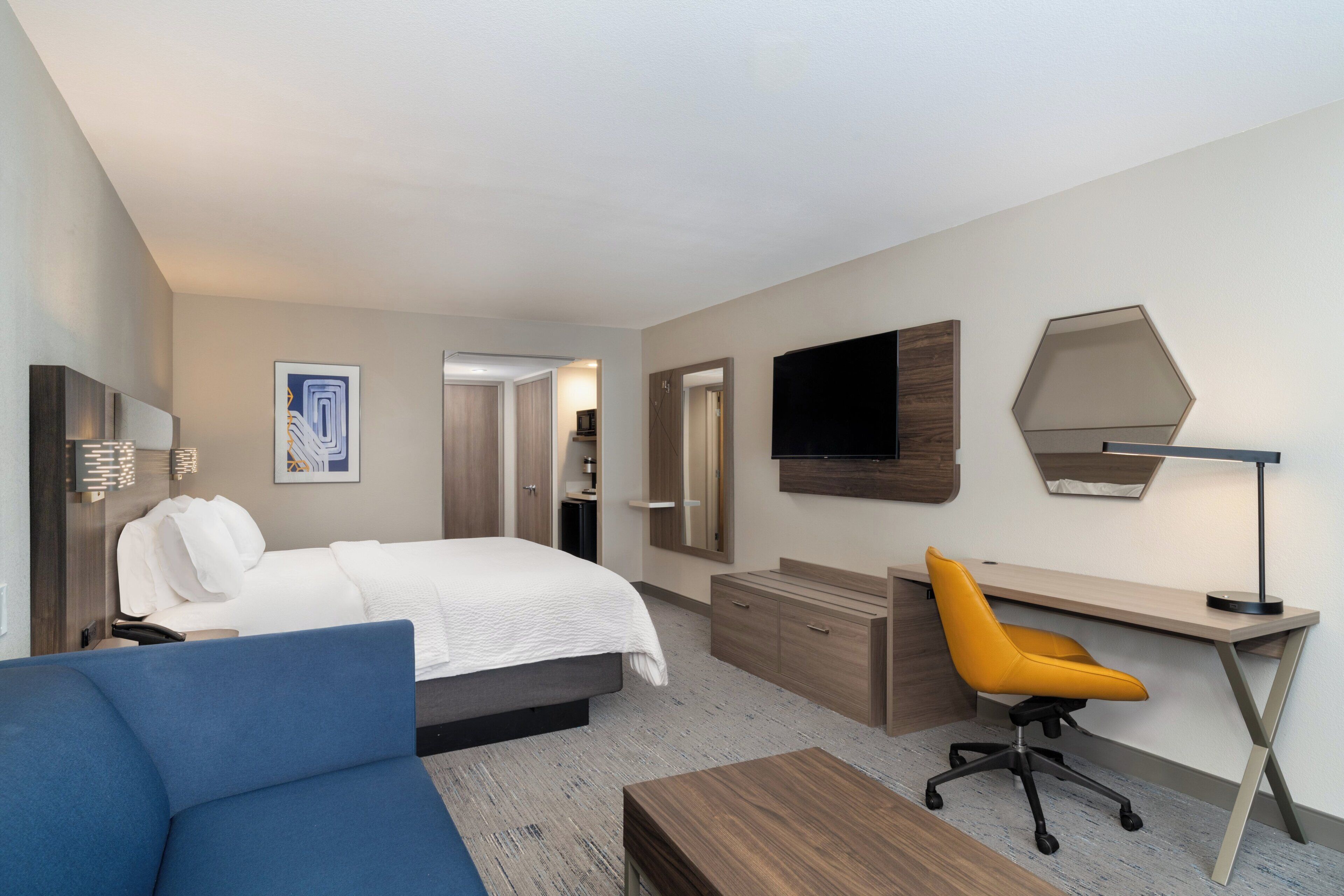 Una habitación de hotel con cama, escritorio, silla, televisor y espejo.