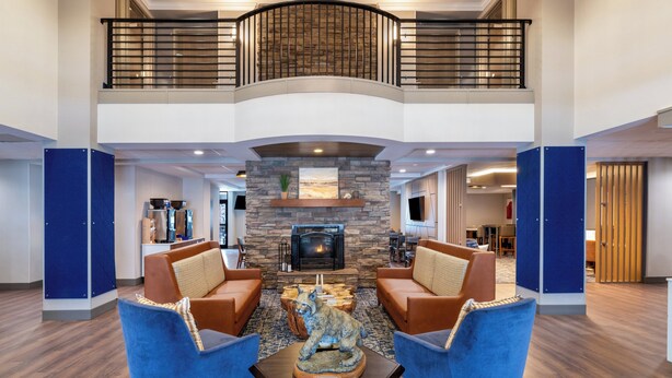 Un hall d’entrée spacieux avec une cheminée, deux fauteuils bleus, une table basse en bois et un plafond haut avec un balcon.