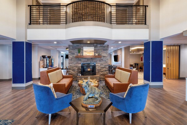 A spacious lobby with a fireplace, two blue armchairs, a wooden coffee table, and a high ceiling with a balcony. - Holiday Inn Express Hotel & Suites Bozeman West by IHG (Bozeman)