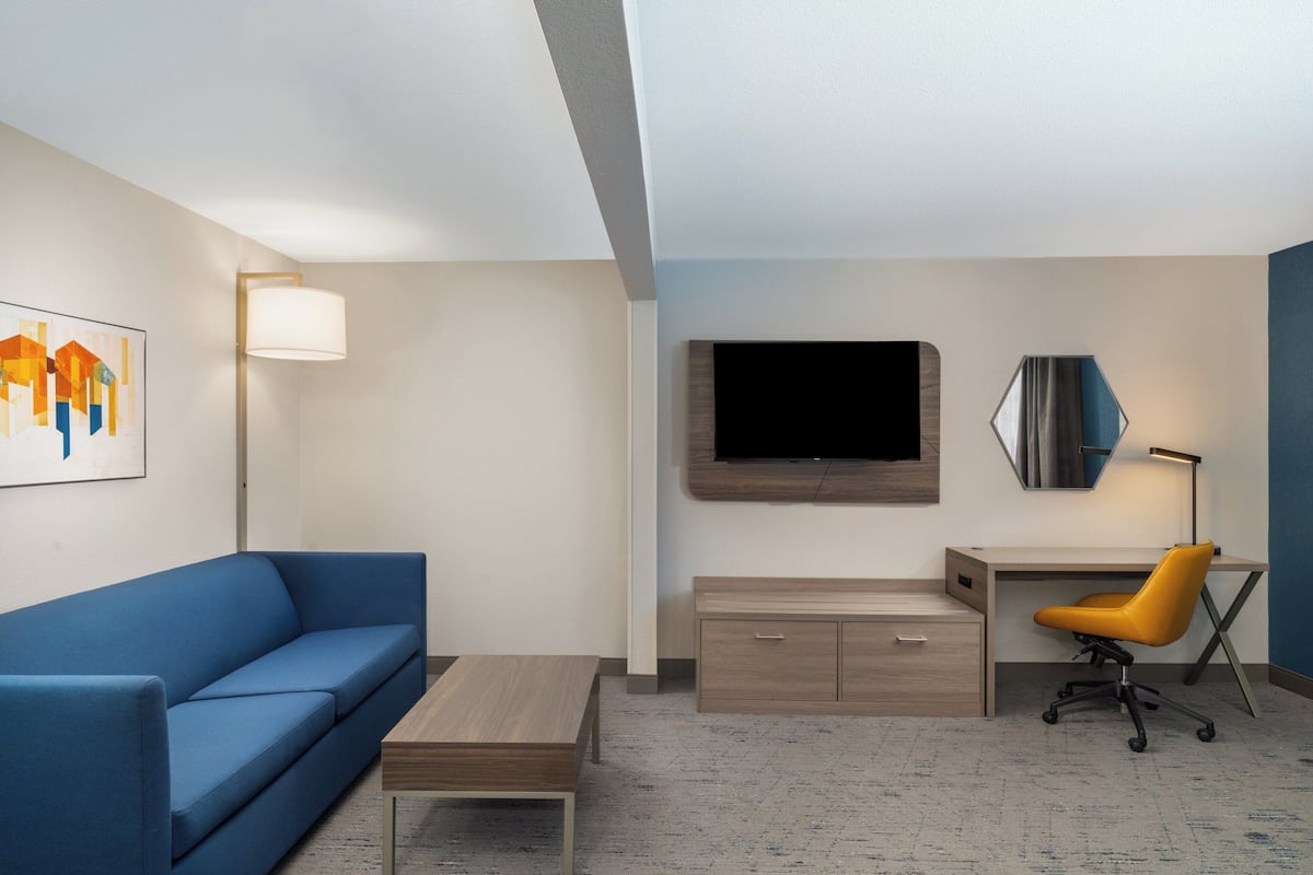 A modern hotel room with a blue sofa, a wooden desk, a yellow office chair, a flat-screen TV, and a painting on the wall.