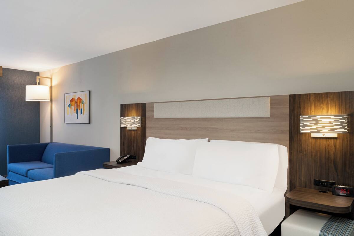 A hotel room with a bed, a blue chair, a nightstand with a phone, and wall-mounted lamps.