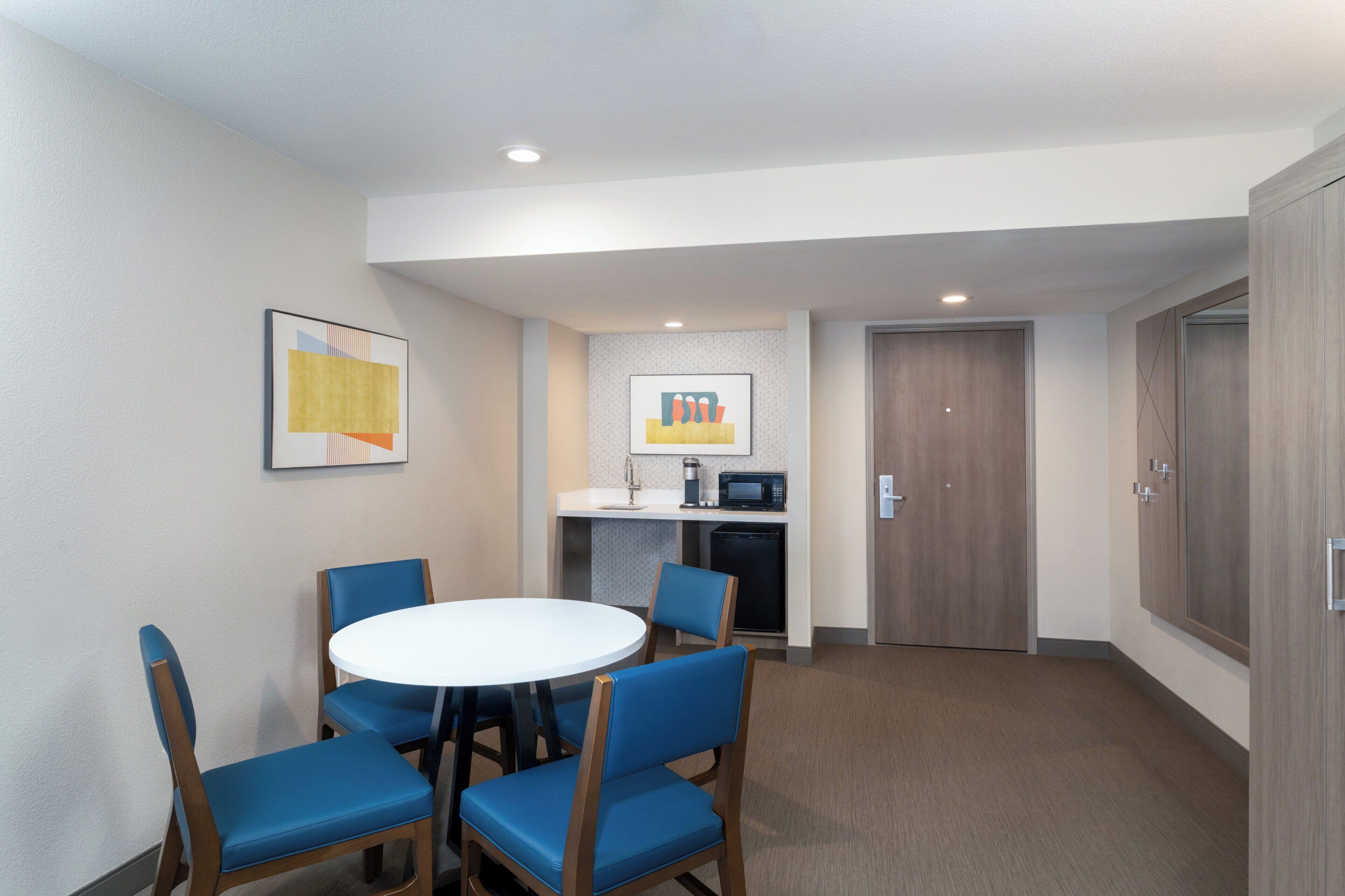 A hotel room with a round table and four chairs, a kitchenette, and a door.