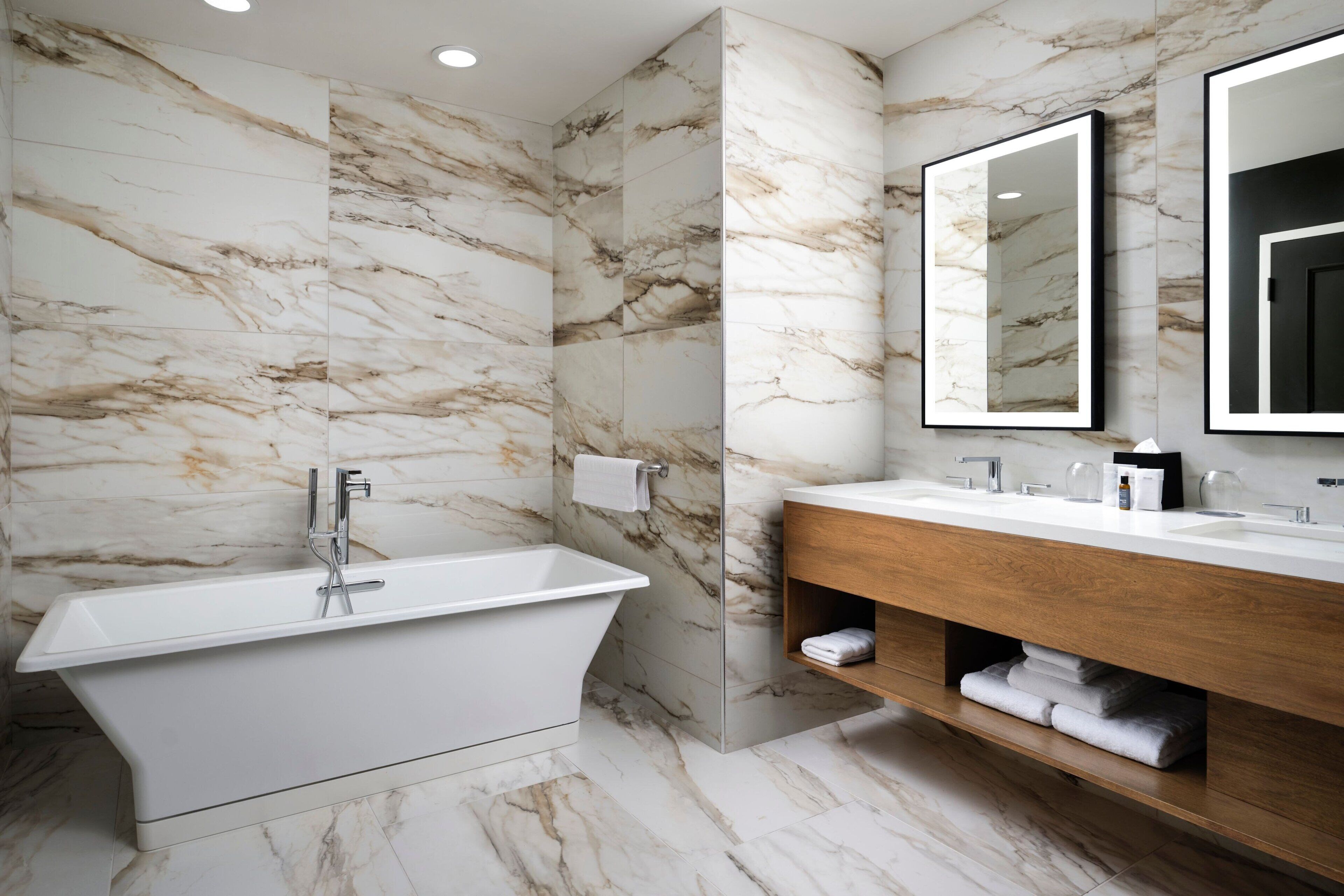A modern bathroom with a freestanding bathtub, a wooden vanity, and marble walls.