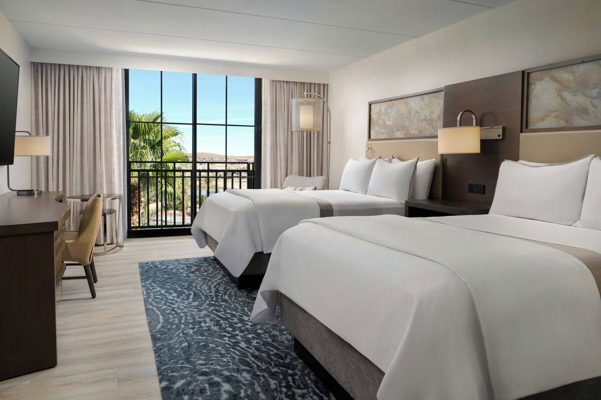A hotel room with two beds, a desk, a chair, a TV, and a balcony with a view.