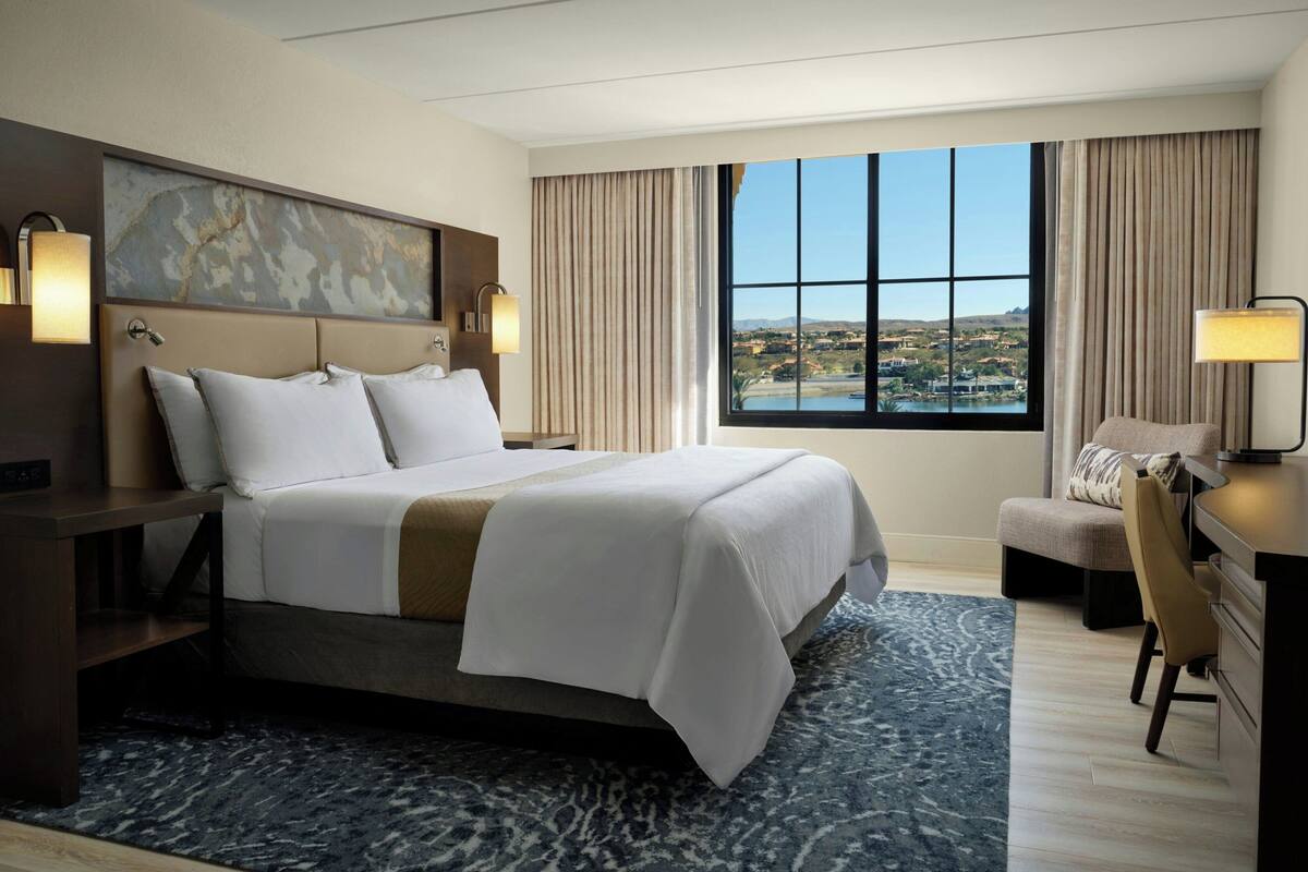 A hotel room with a large bed, a desk, a chair, and a view of a landscape through the window.