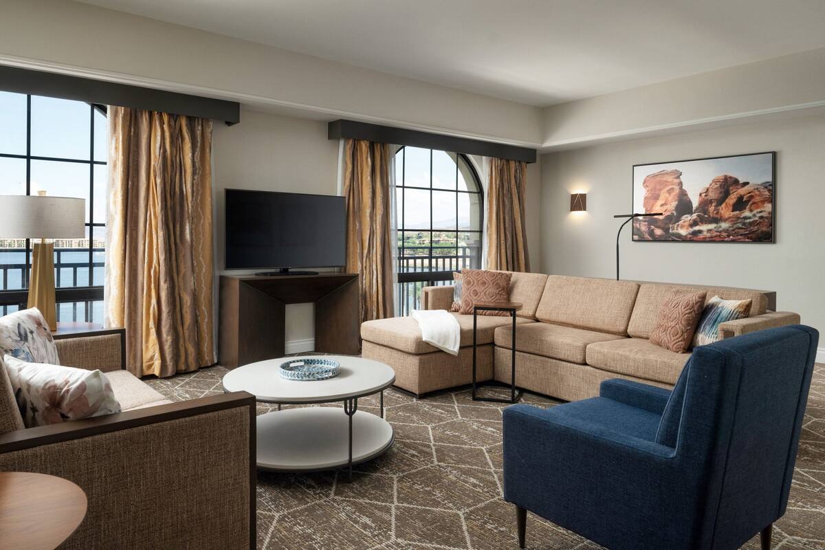 A modern hotel room with a sectional sofa, a blue armchair, a round coffee table, and a flat-screen TV.