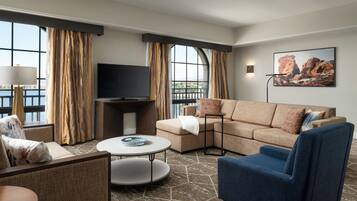 A modern hotel room with a sectional sofa, a blue armchair, a round coffee table, and a flat-screen TV.