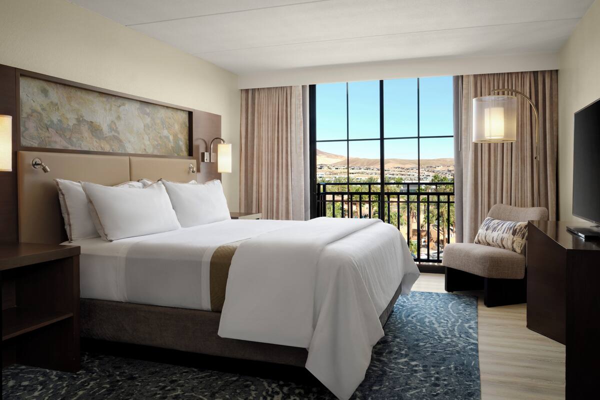 A hotel room with a large bed, a desk, a chair, a TV, a balcony with a view, and a painting on the wall.