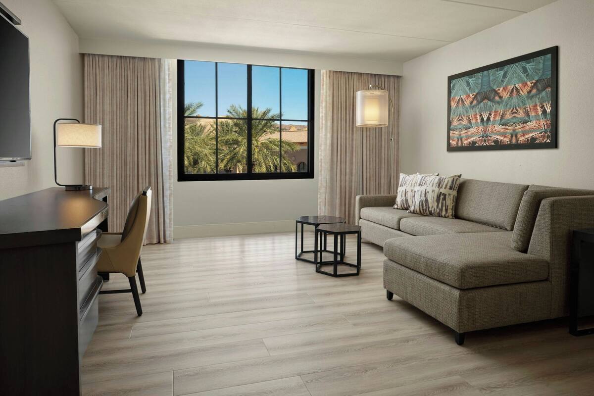 A hotel room with a large window, a sofa, a desk with a chair, and a small table.