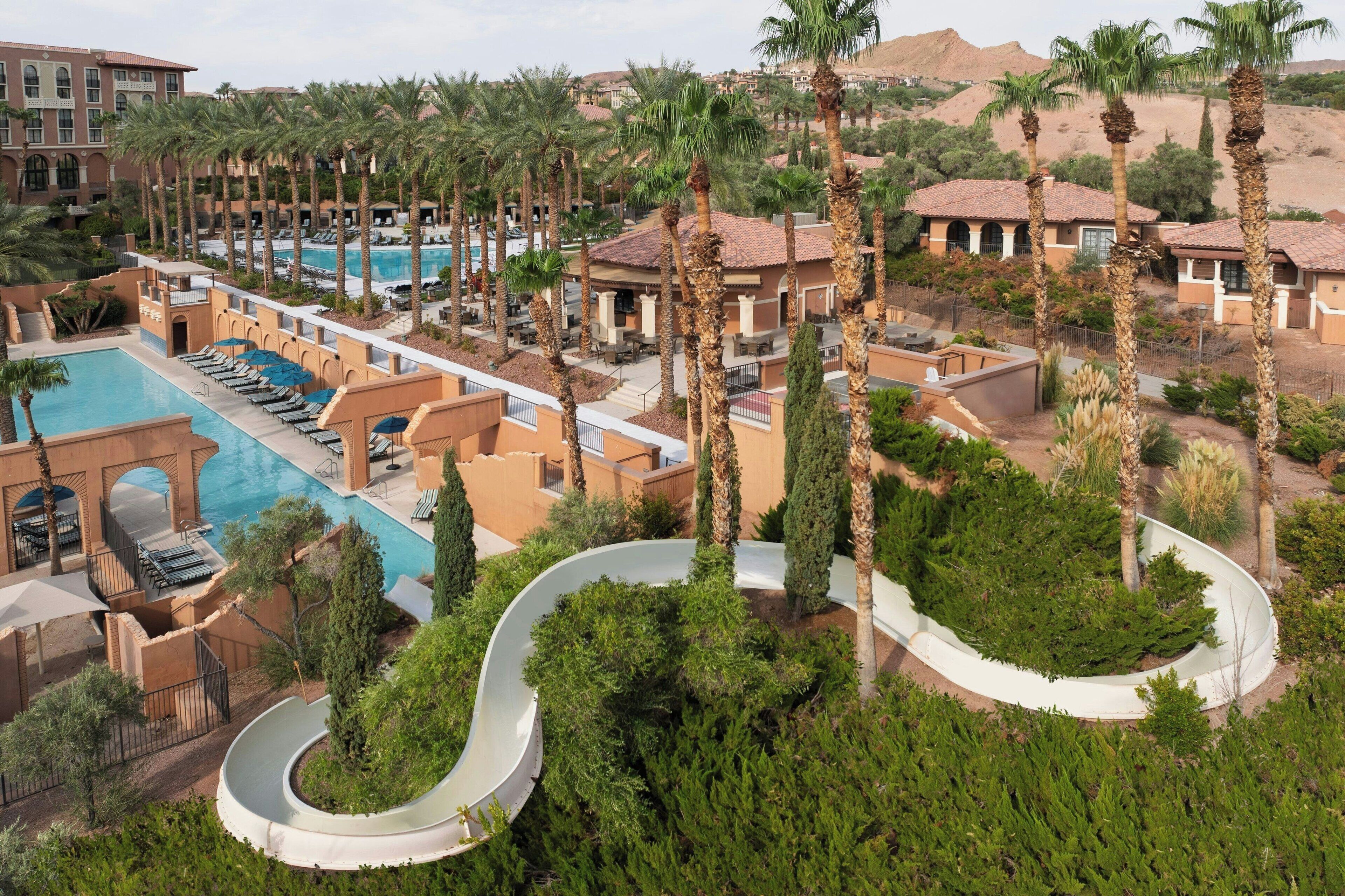 A resort with a swimming pool, lounge chairs, palm trees, and a winding water slide.