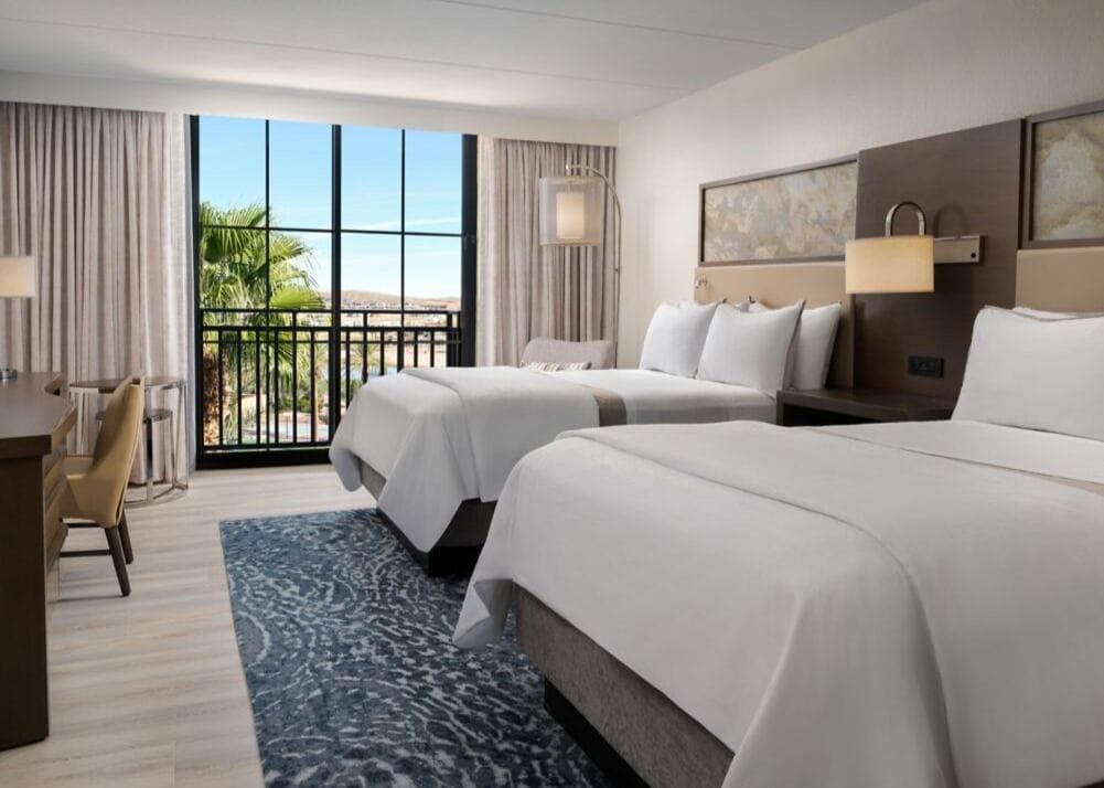 A hotel room with two beds, a desk, and a view of the outdoors.