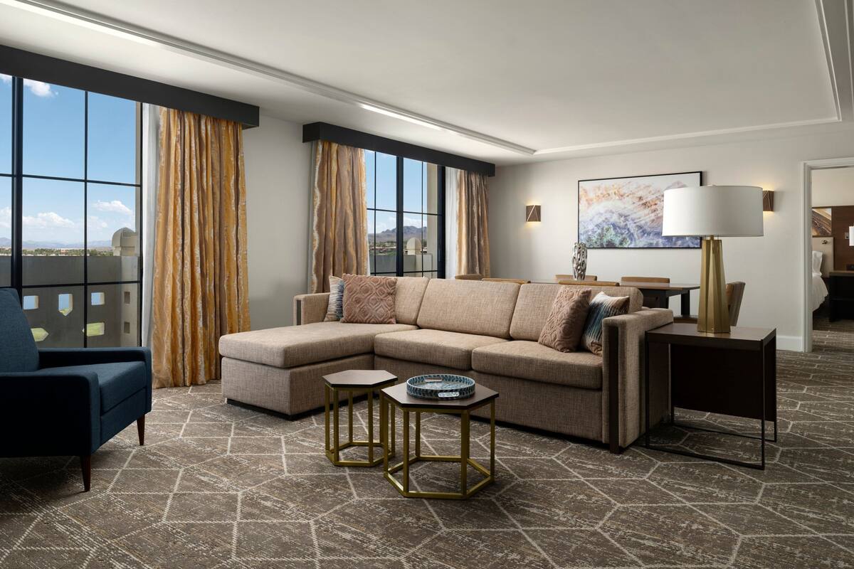 A modern living room with a sectional sofa, two side tables, a lamp, and a view of the city through large windows.