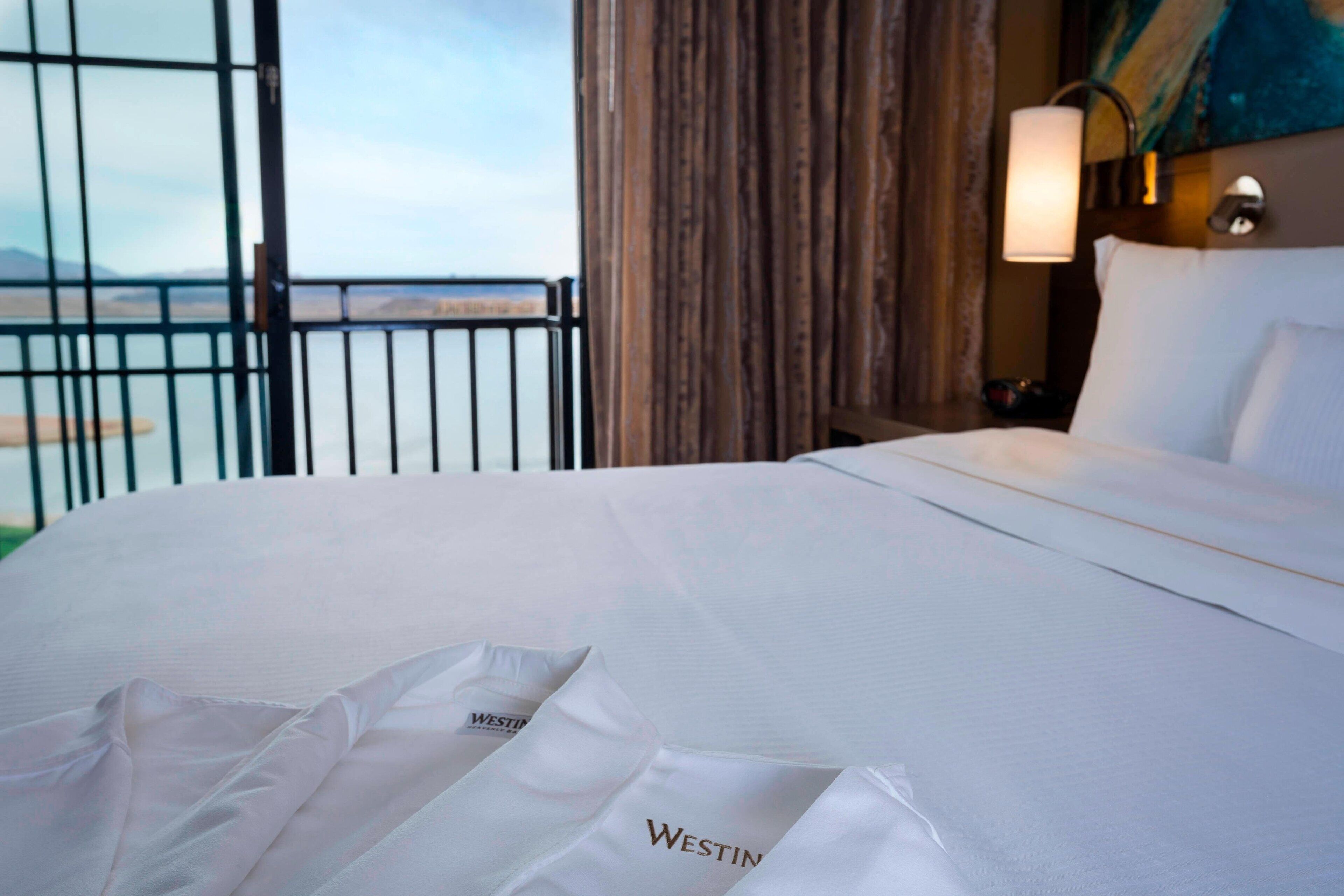 A hotel room with a large bed, a view of the sea through a balcony, and a Westin logo on the bedspread.