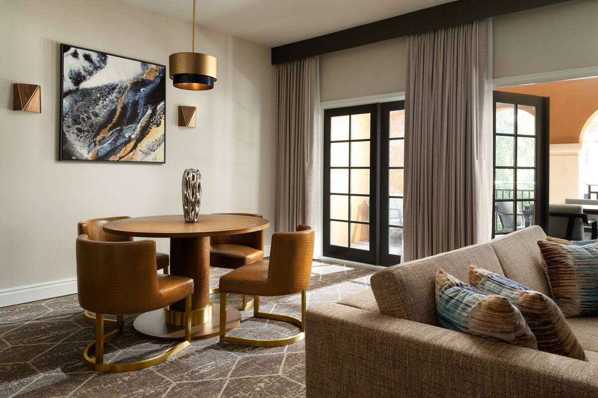 A modern living room with a round dining table, leather chairs, a sofa with patterned cushions, and a large abstract painting on the wall.