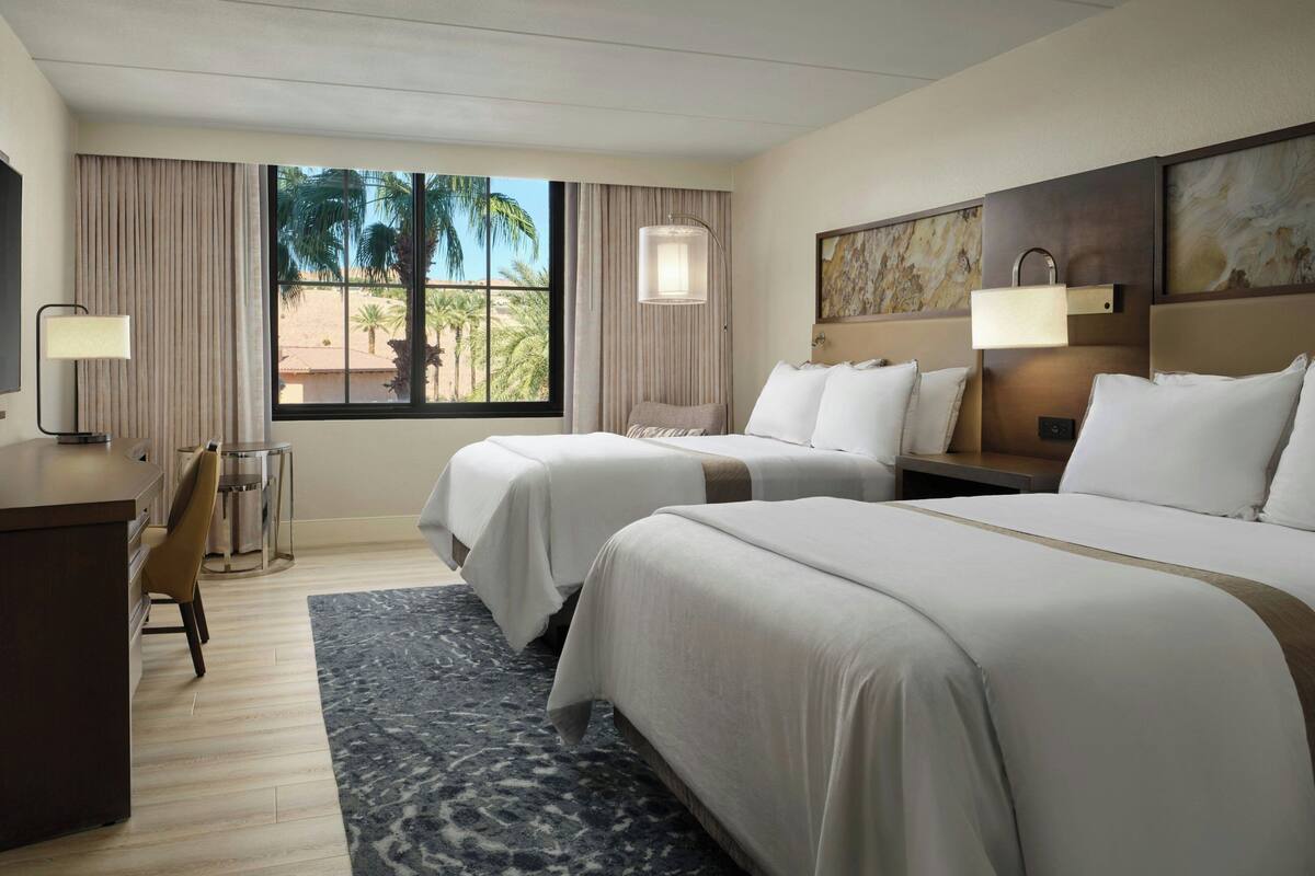 A hotel room with two beds, a desk, a chair, a small table, and a window with curtains.