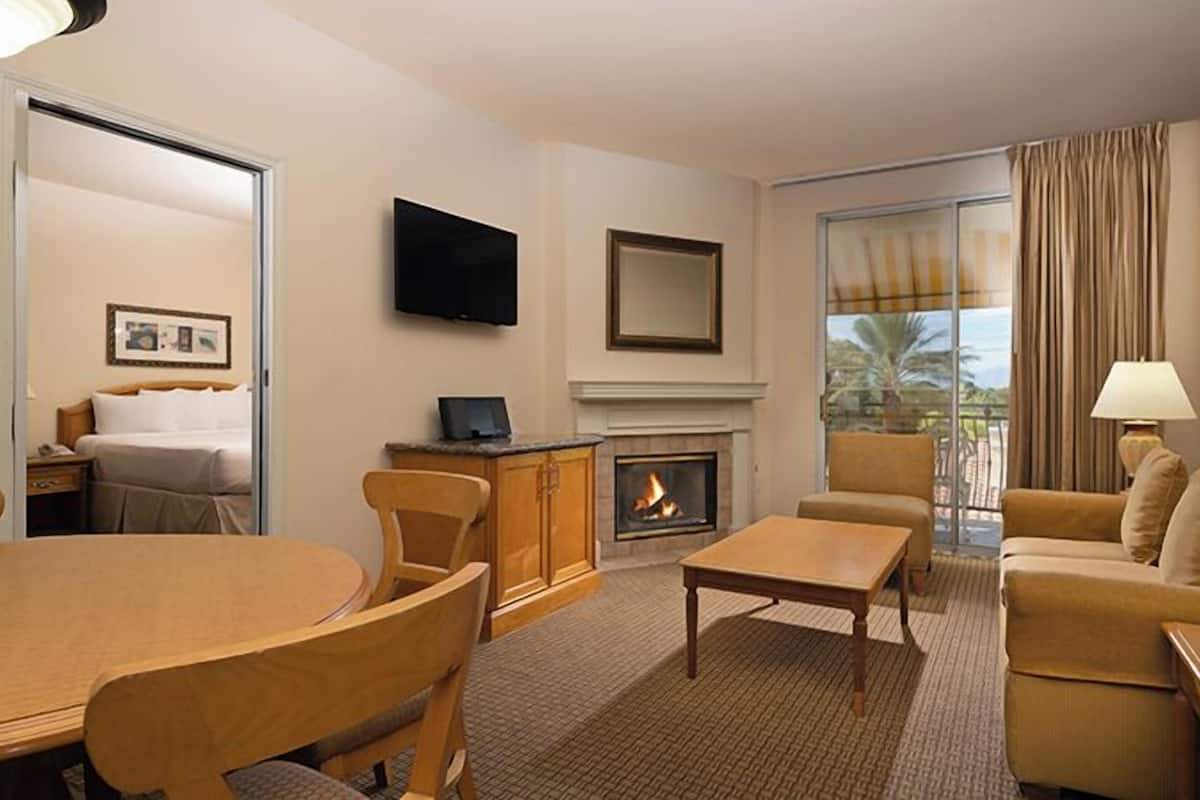 A hotel room with a dining table, chairs, a sofa, a fireplace, a TV, and a large window with curtains.