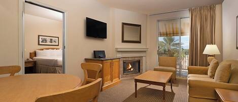 Habitación de hotel con una mesa de comedor, sillas, un sofá, una chimenea, un televisor y un ventanal con cortinas.