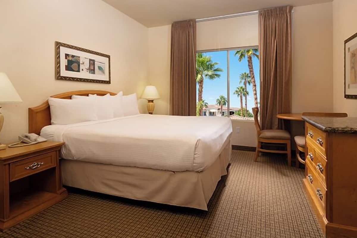 A hotel room with a large bed, two bedside lamps, a wooden nightstand, a dresser, and a window with a view of palm trees.