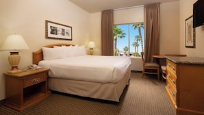 Habitación de hotel con una cama grande, dos lámparas de noche, una mesita de noche de madera, un aparador y una ventana con vistas a palmeras.