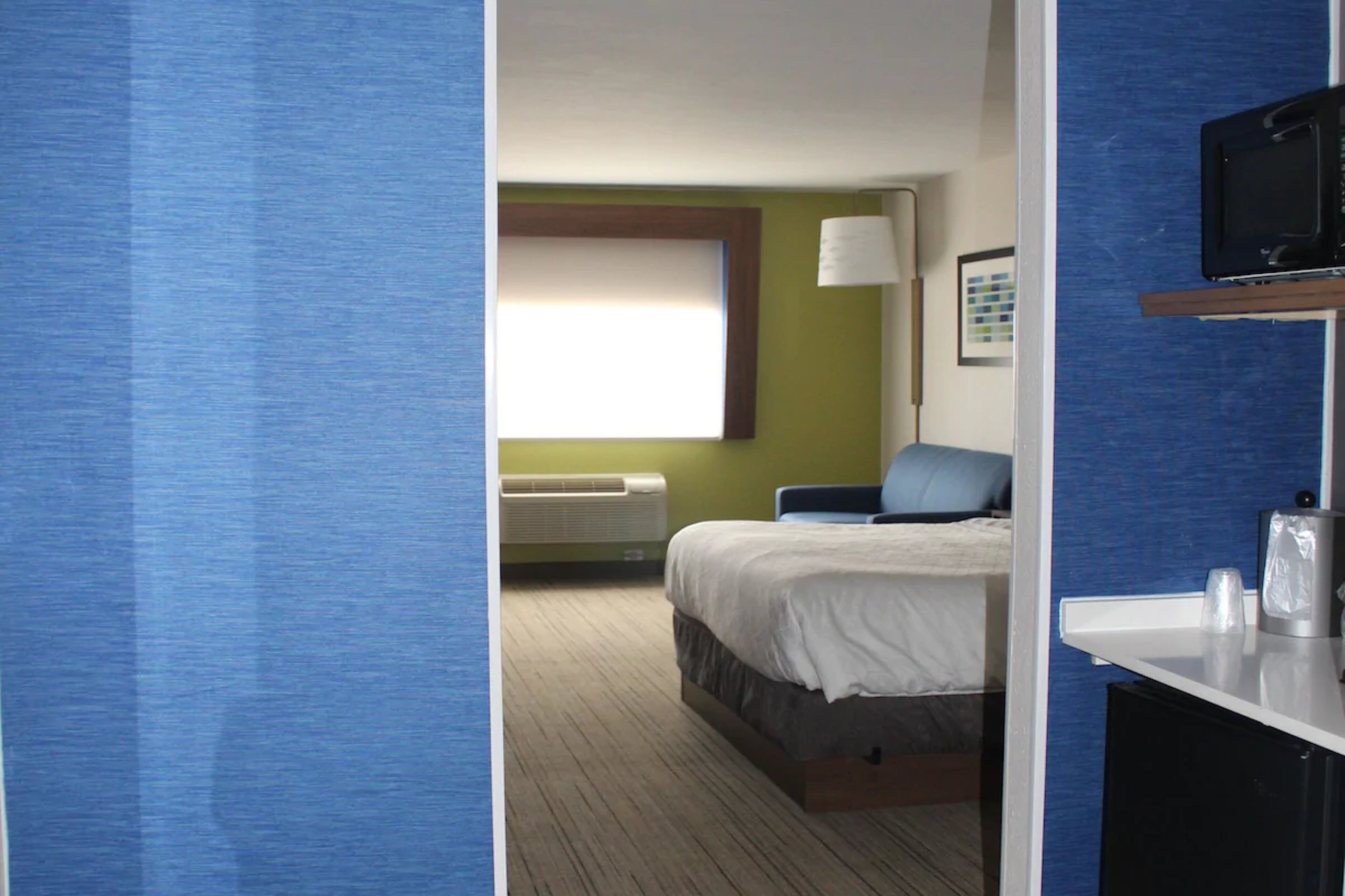 A hotel room with a bed, a desk with a chair, a television, and a window with blinds.