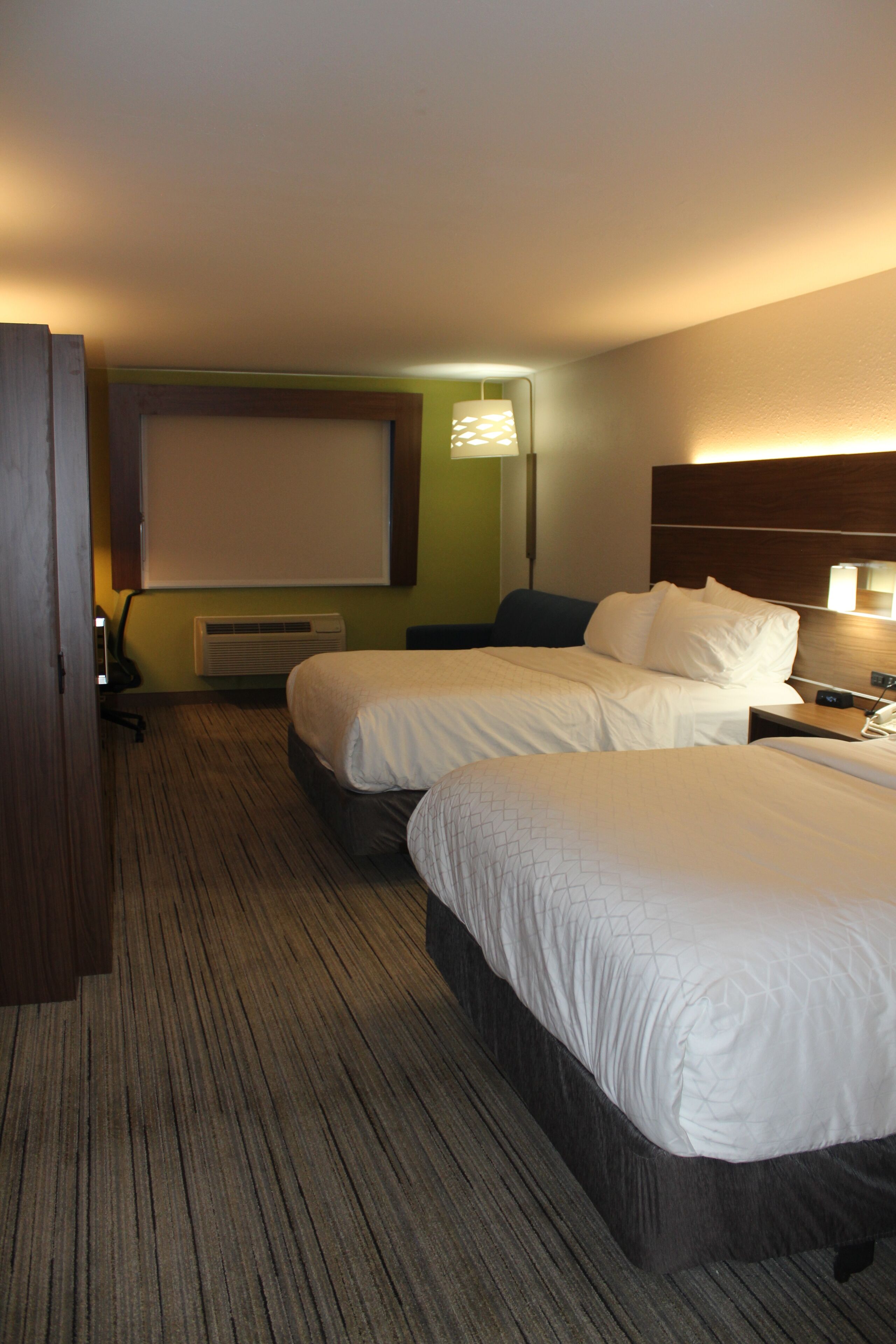 A hotel room with two beds, a desk, a chair, a projector screen, and an air conditioning unit.