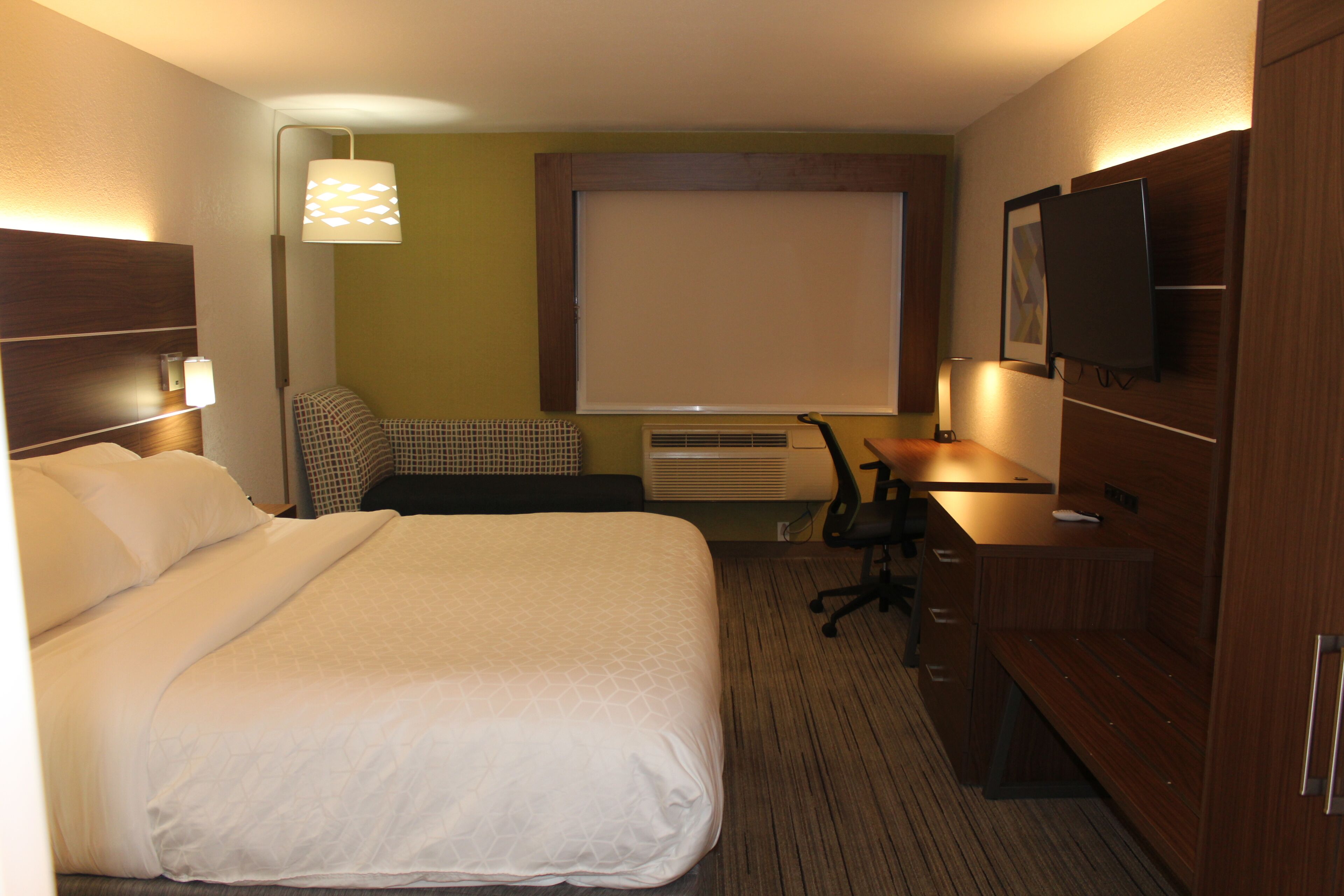 A hotel room with a large bed, a desk, a chair, a television, and a window with blinds.