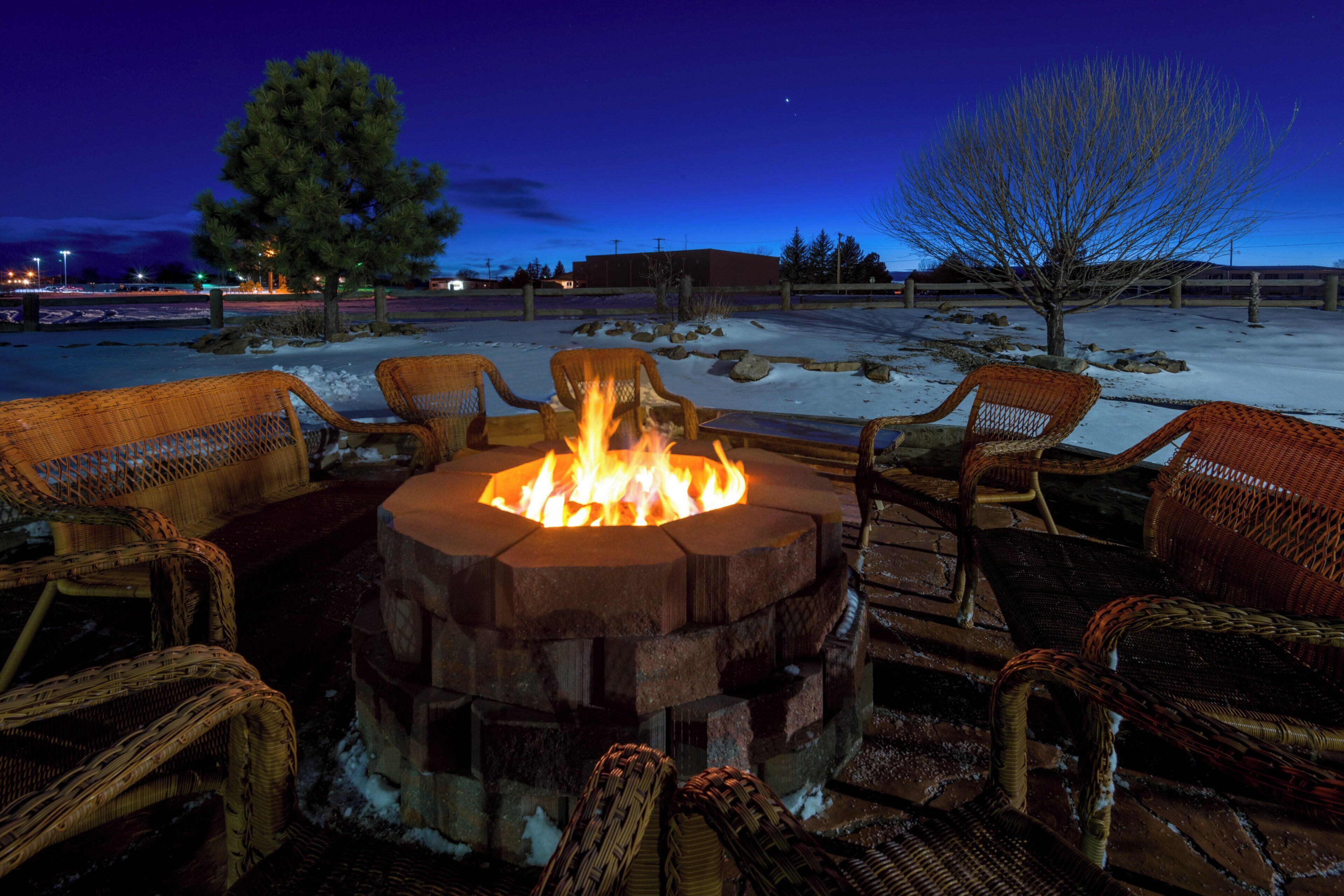 A fire pit surrounded by wicker chairs in a snowy outdoor setting at dusk.