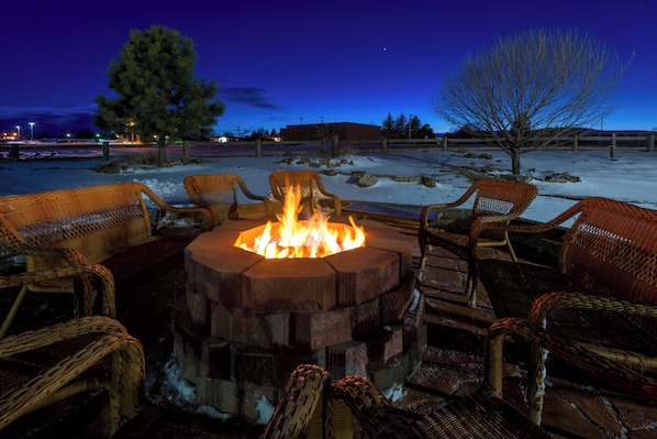 A fire pit surrounded by wicker chairs in a snowy outdoor setting at dusk.