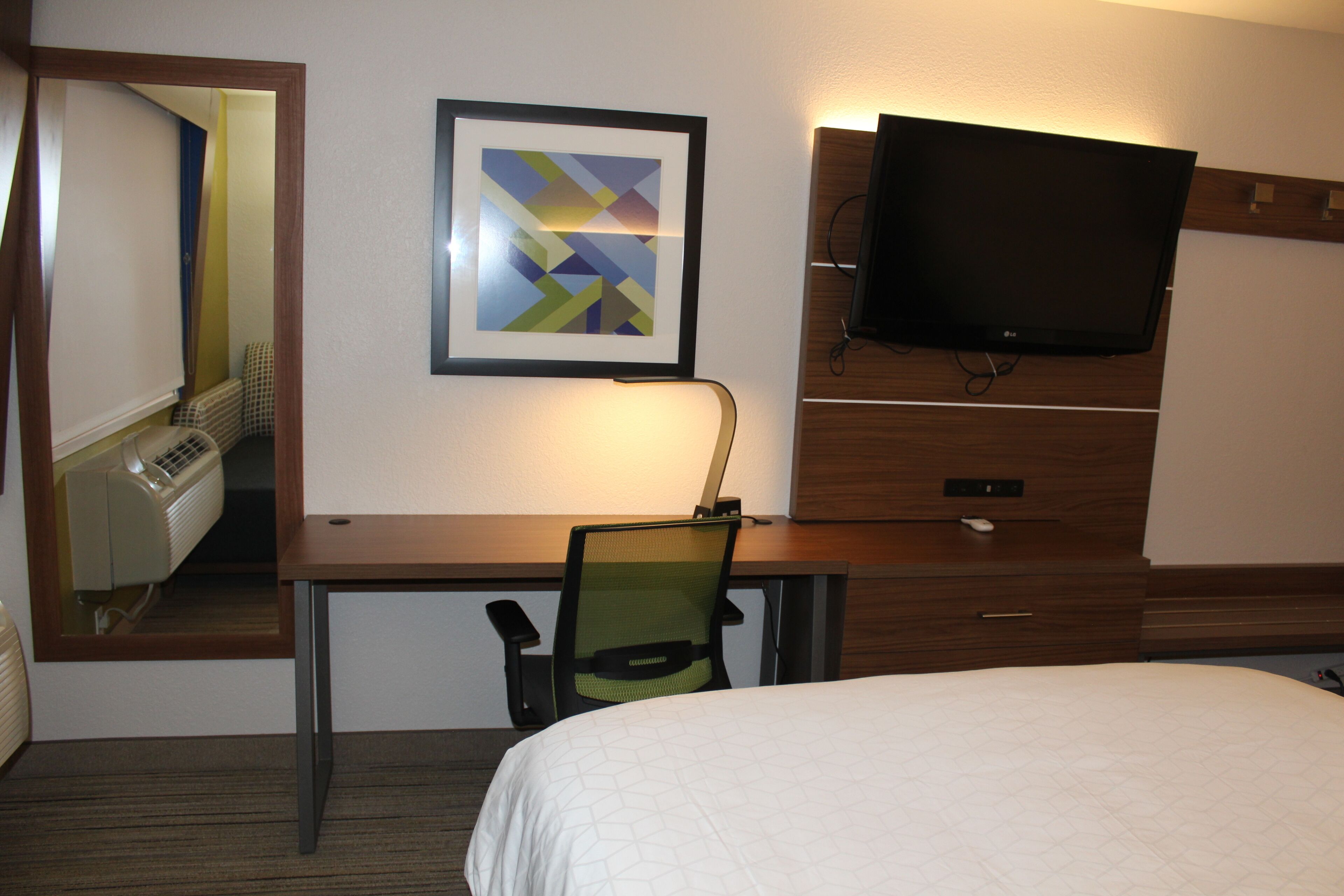 A hotel room with a bed, a desk with a chair, a television mounted on the wall, and a framed picture.