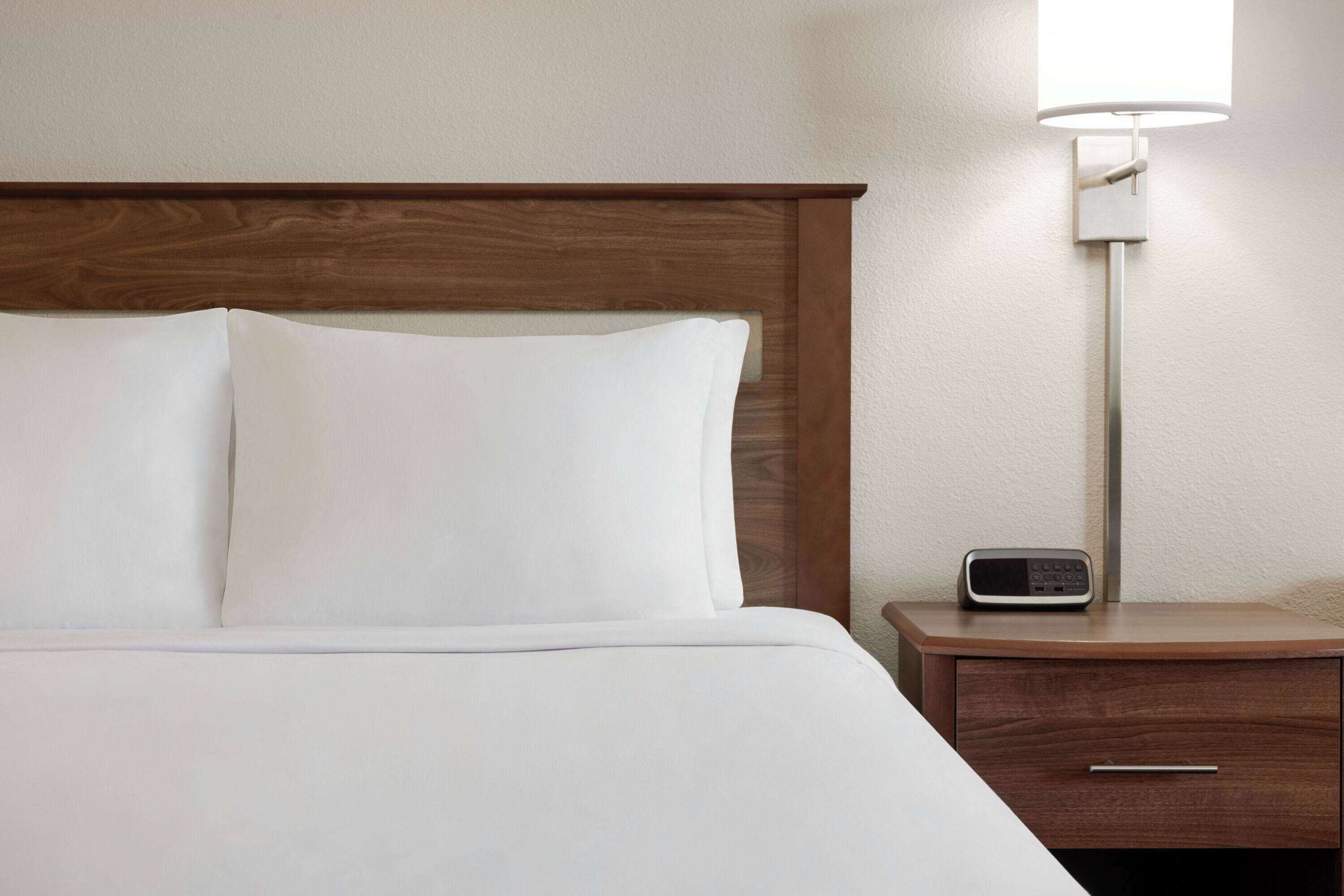 Una cama bien hecha con sábanas blancas y un cabecero de madera, una mesita de noche con una lámpara y un pequeño dispositivo electrónico sobre la mesa.