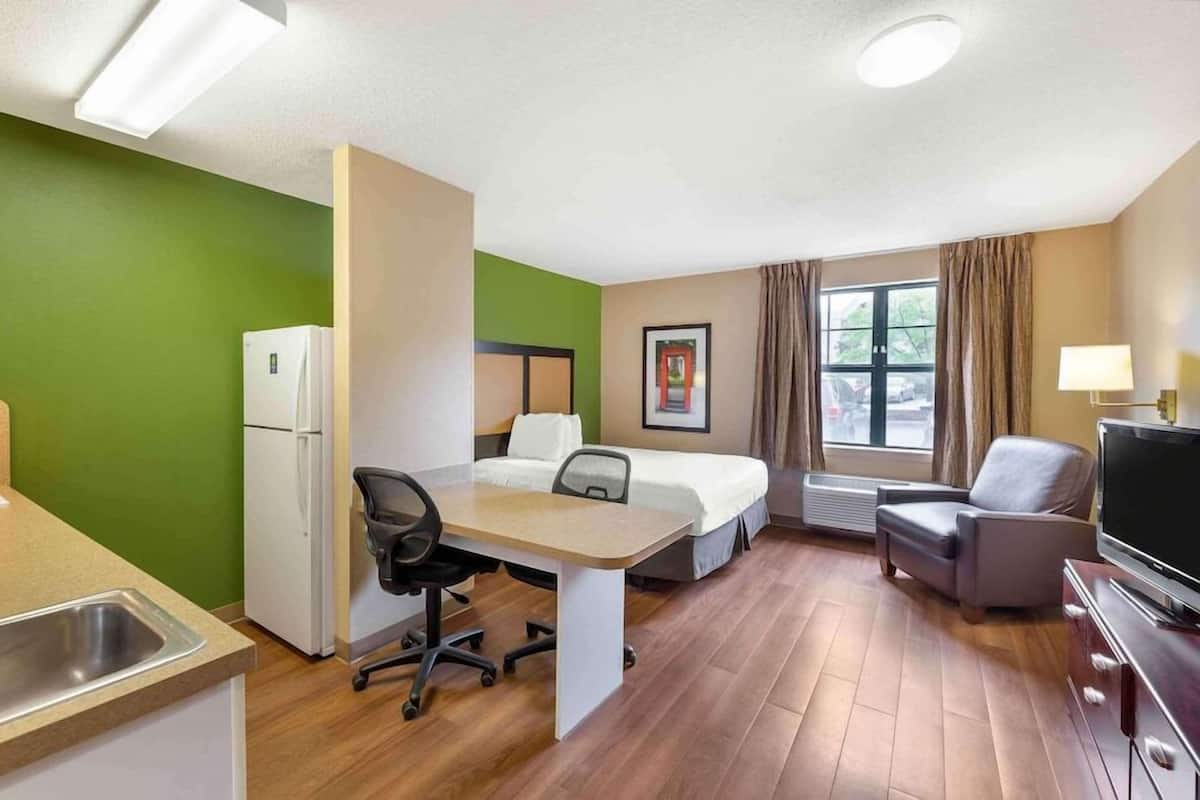 A hotel room with a kitchenette, a bed, a desk, a chair, a TV, and a window with curtains.