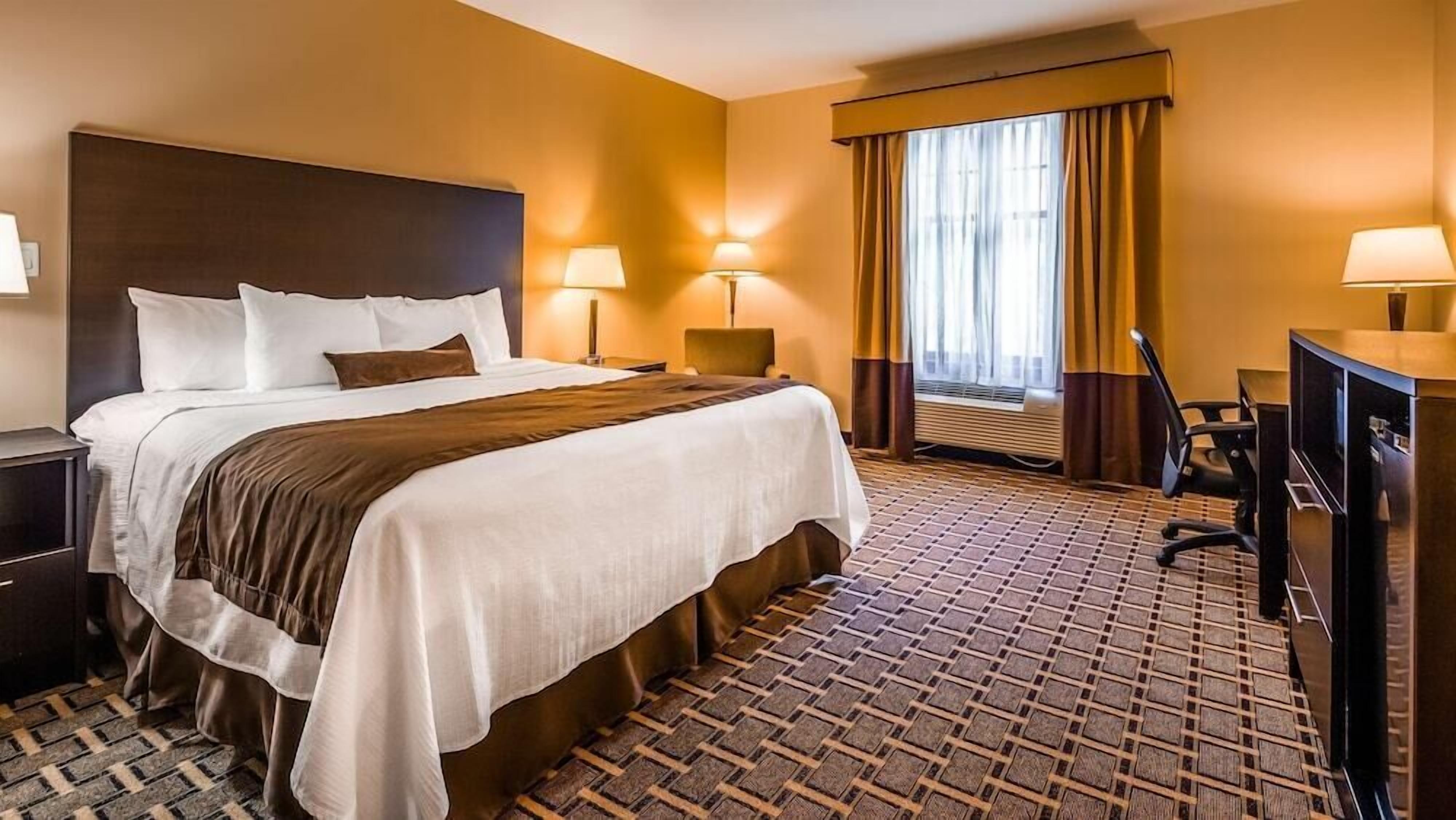 A hotel room with a large bed, two bedside lamps, a desk with a chair, a television, and a window with curtains.