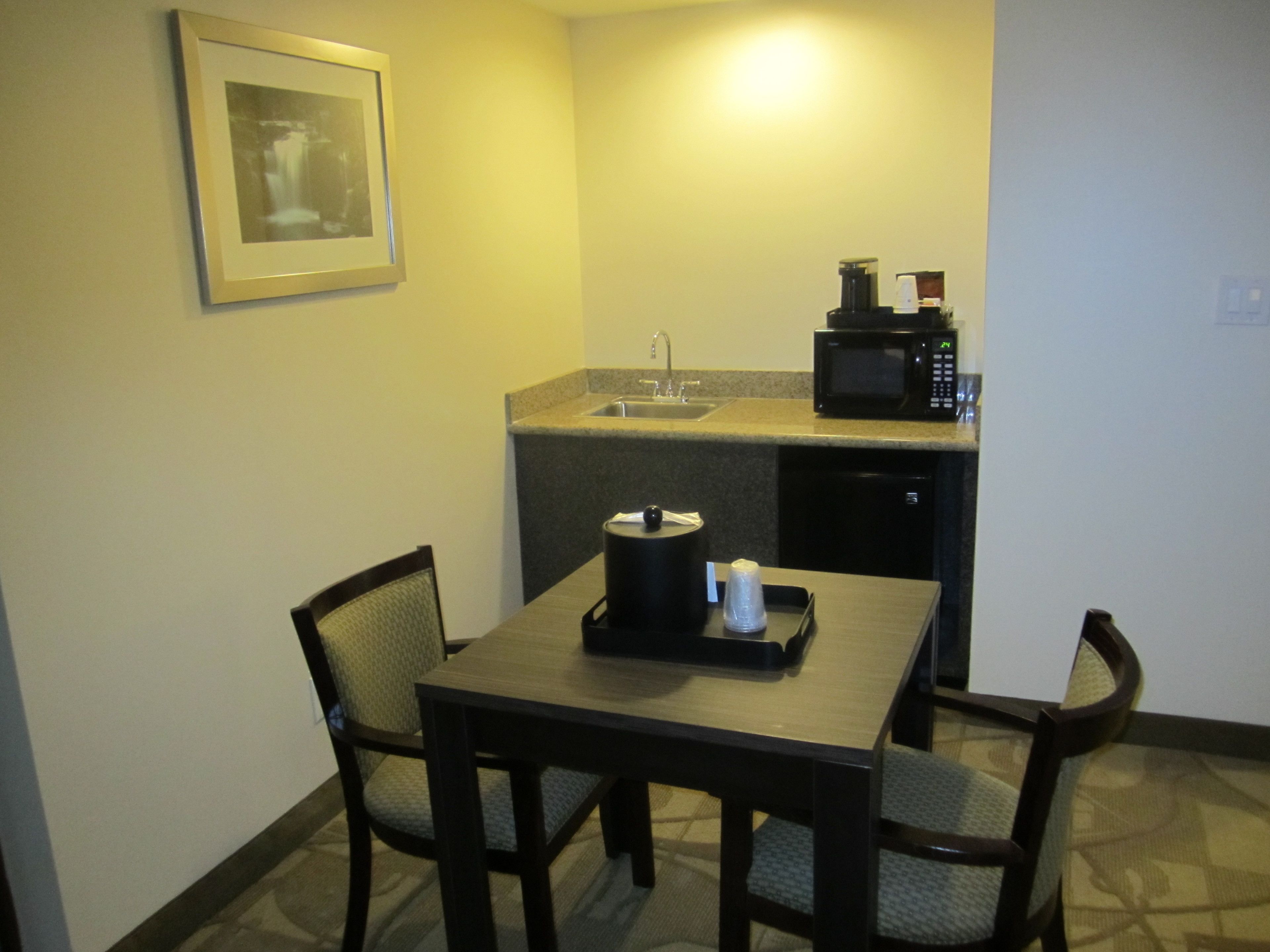 Une petite cuisine équipée d’un four à micro-ondes, d’un évier et d’un tableau encadré au mur. On aperçoit une table avec des chaises au premier plan.