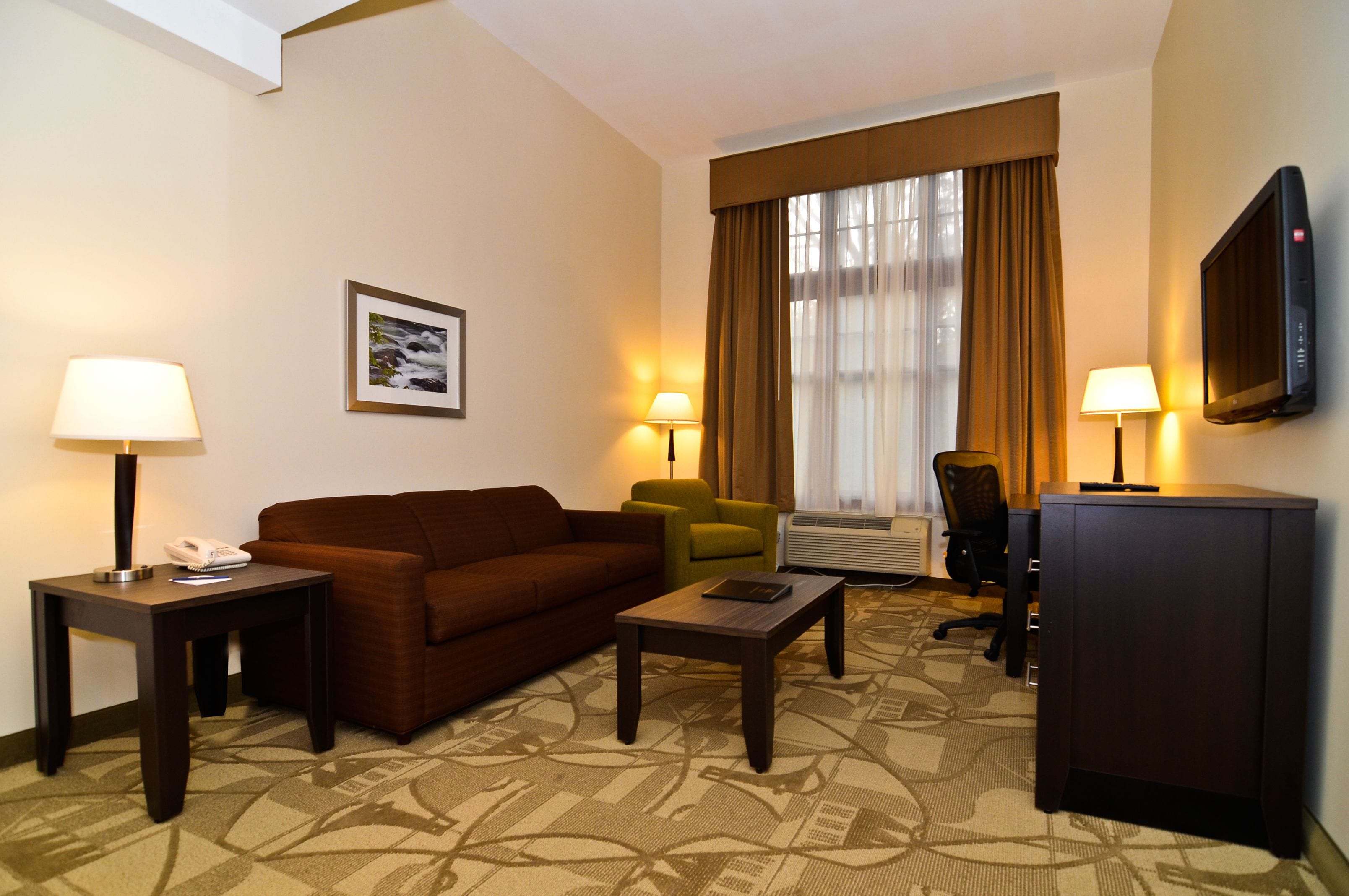 Ein Hotelzimmer mit einem braunen Sofa, zwei Sesseln, einem Couchtisch, einem Fernseher und einem gerahmten Bild an der Wand.