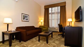 Une chambre d’hôtel avec un canapé marron, deux fauteuils, une table basse, un téléviseur et un tableau encadré au mur.