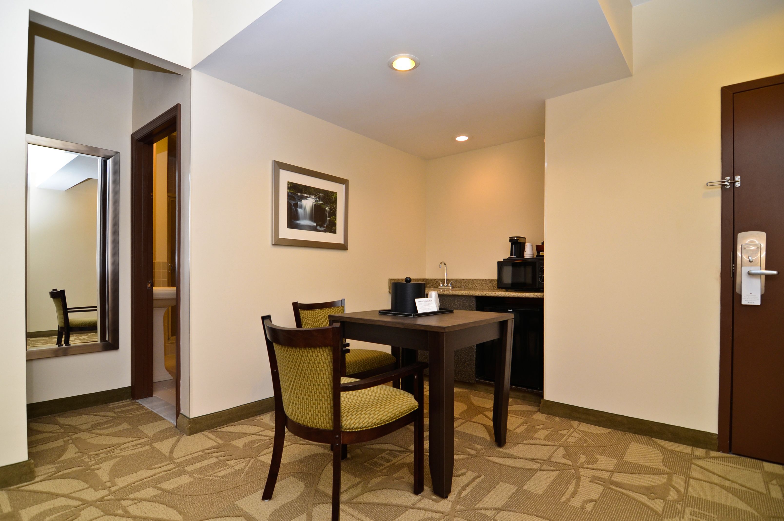 A hotel room with a dining table, two chairs, a framed picture on the wall, a kitchen area, and a door with a lock.