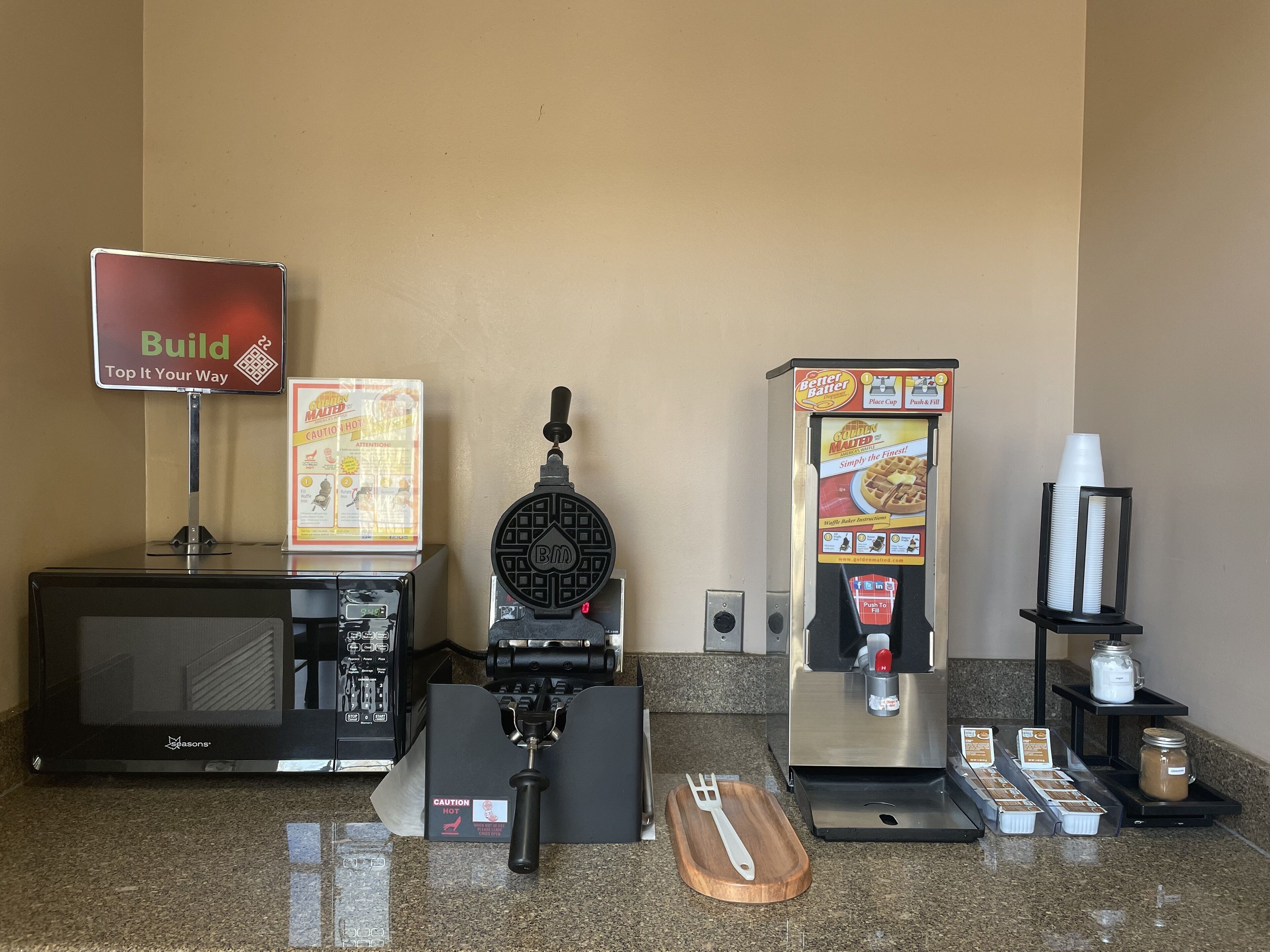 A self-service breakfast station with a waffle maker, coffee machine, and condiment dispensers.