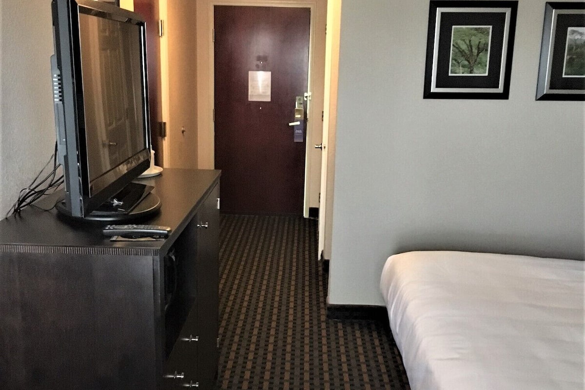 A hotel room with a bed, a dresser with a television, a door, and two framed pictures on the wall.