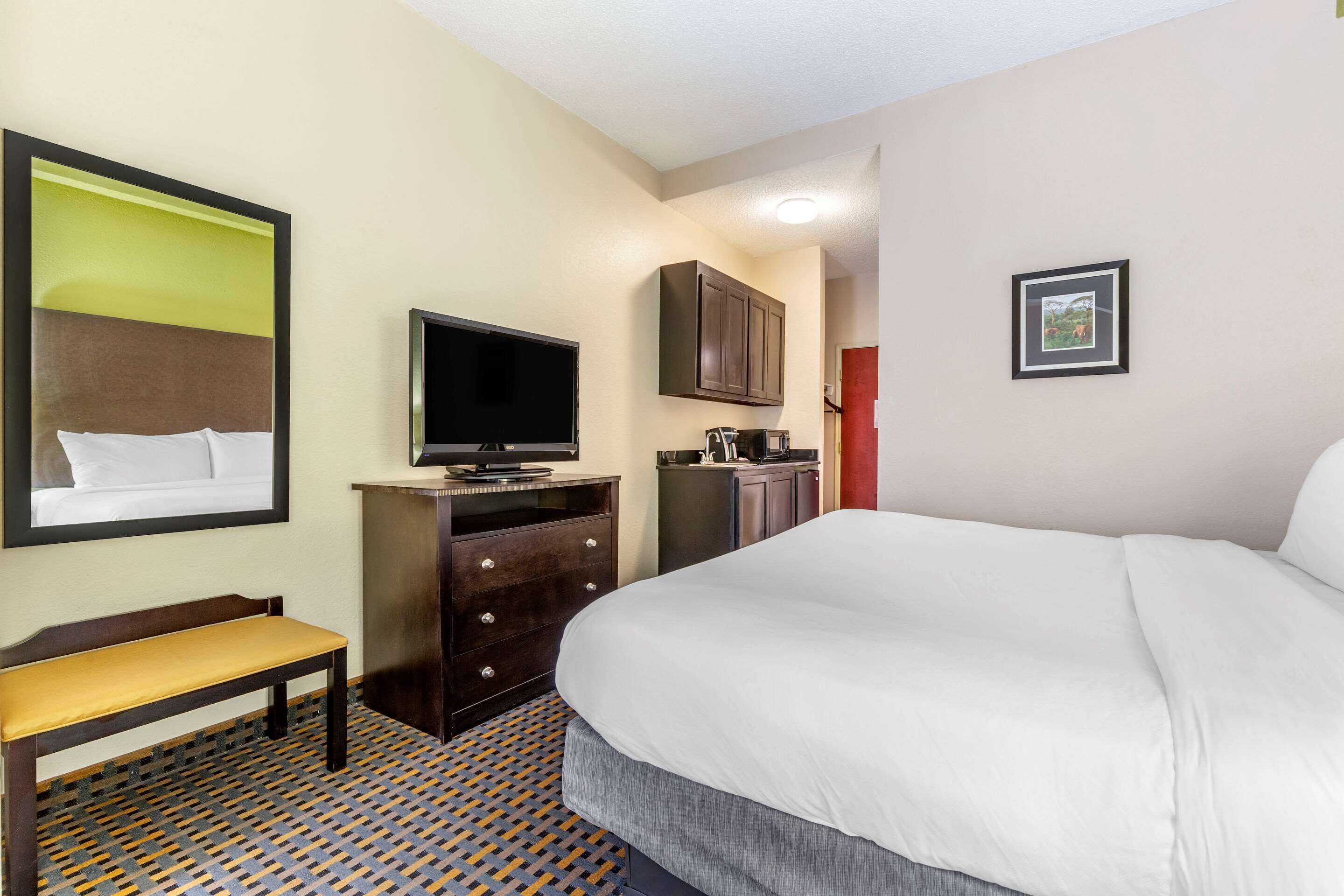 Una habitación de hotel con una cama grande, un televisor en una cómoda, un banco, una pequeña cocina y un cuadro en la pared.
