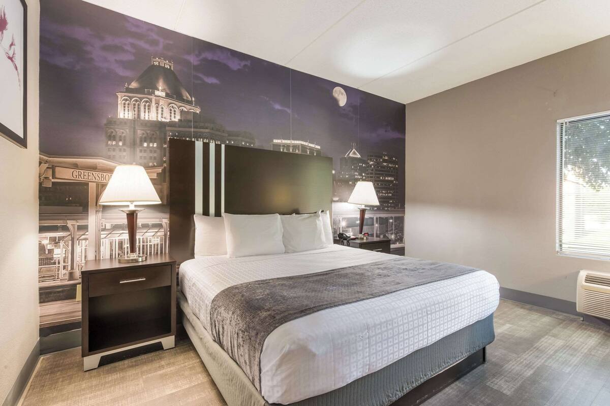 Une chambre d’hôtel avec un grand lit, deux tables de chevet avec des lampes, une petite table et une fresque représentant un paysage urbain sur le mur.