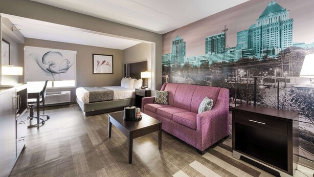 Une chambre d’hôtel avec un lit, un bureau, un canapé rose, une table basse et une fresque représentant un paysage urbain sur le mur.