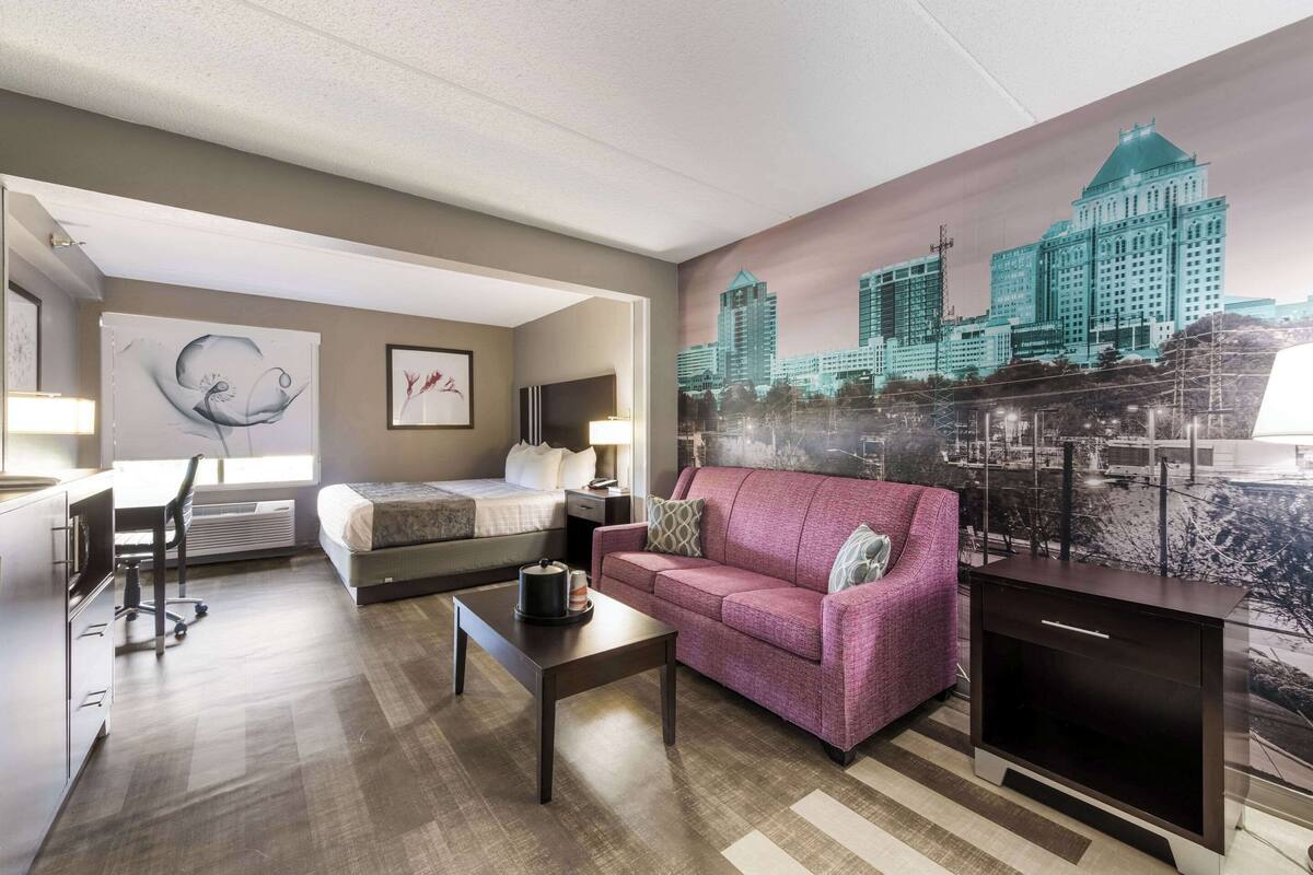 Une chambre d’hôtel avec un lit, un bureau, un canapé rose, une table basse et une fresque représentant un paysage urbain sur le mur.
